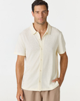 A man wearing a short-sleeve knit polo shirt in cream and tan trousers, standing against a plain light gray studio background.