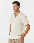 A man wearing a short-sleeve knit polo shirt in a light cream color, paired with beige trousers, photographed against a plain white studio background.