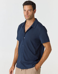 A man wearing a navy blue short-sleeve button-up shirt and beige trousers, standing against a light gray studio background.