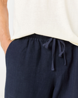 Close-up of a person wearing dark navy drawstring trousers and an off-white top, with one hand in a pocket against a plain light background.