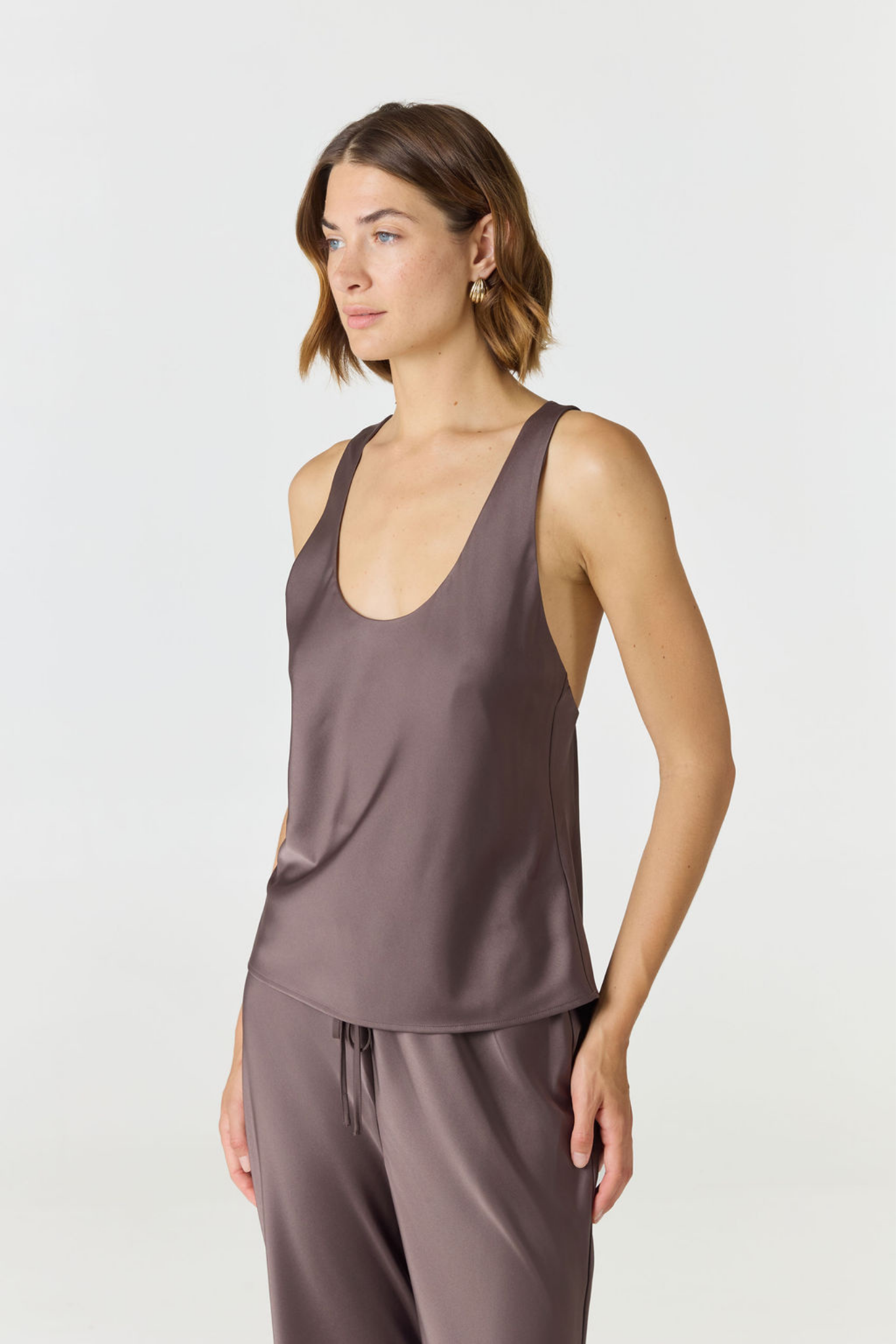 A woman modeling a sleeveless satin-like top and matching drawstring pants in a dusty mauve-brown color, photographed against a clean light gray studio background.