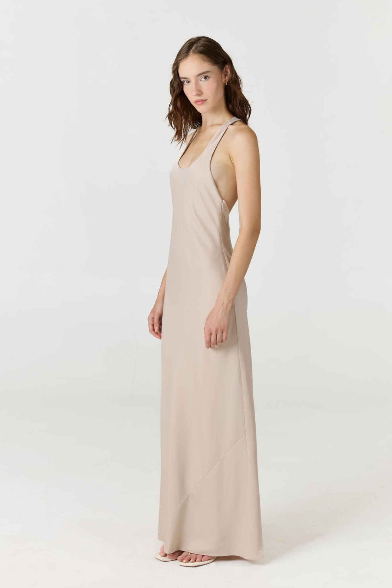A woman modeling a sleeveless, floor-length slip dress in a light beige or nude color, standing against a plain white studio background.
