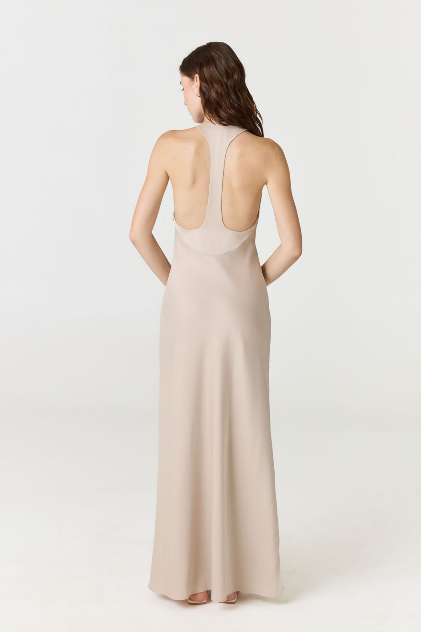 A woman viewed from the back wearing a sleeveless, floor-length nude-beige gown with a wide open back, standing against a clean light gray studio background.