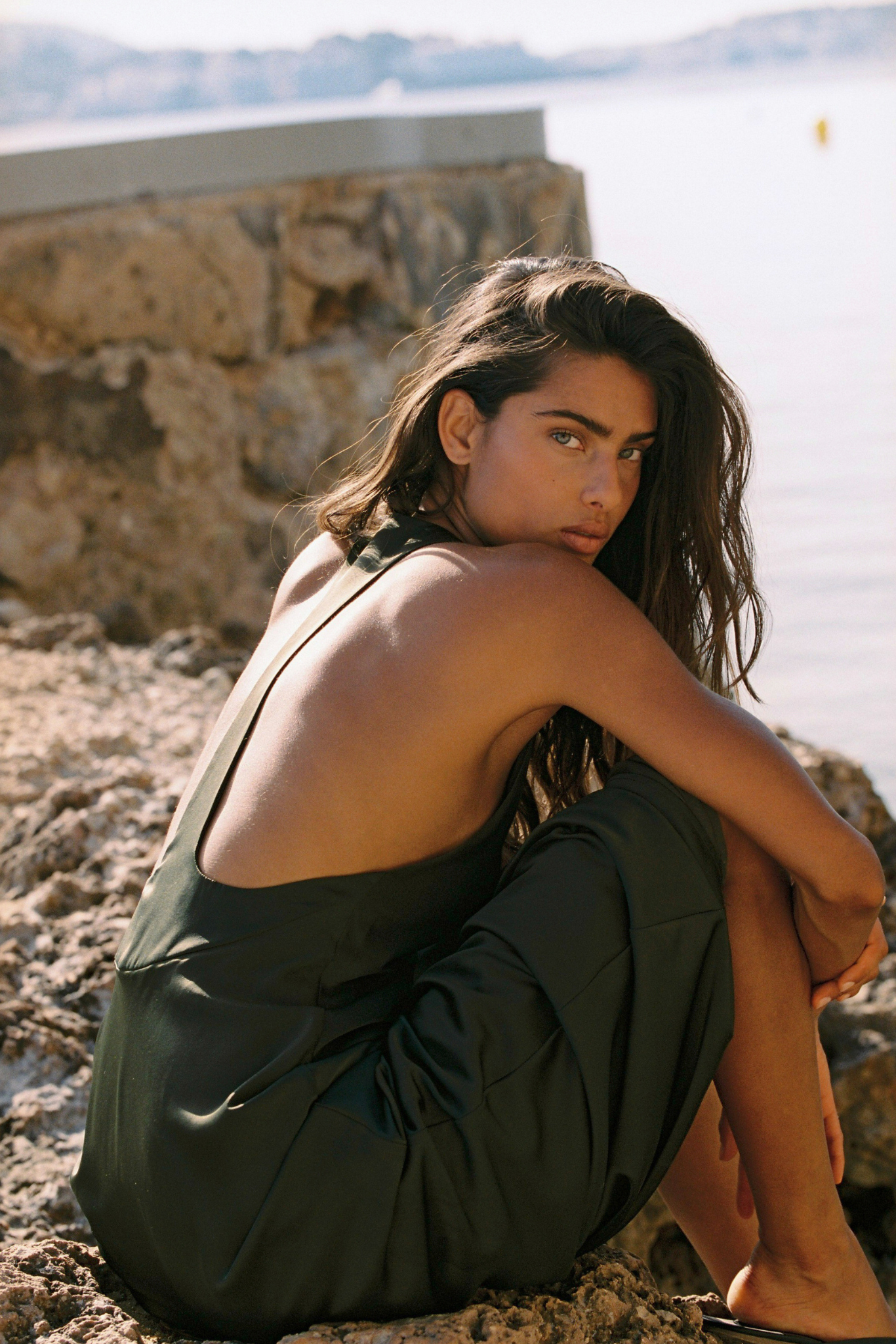 A woman crouches on a rocky shoreline with water and a stone wall in the background, wearing a dark olive green halter-neck dress or lounge set.