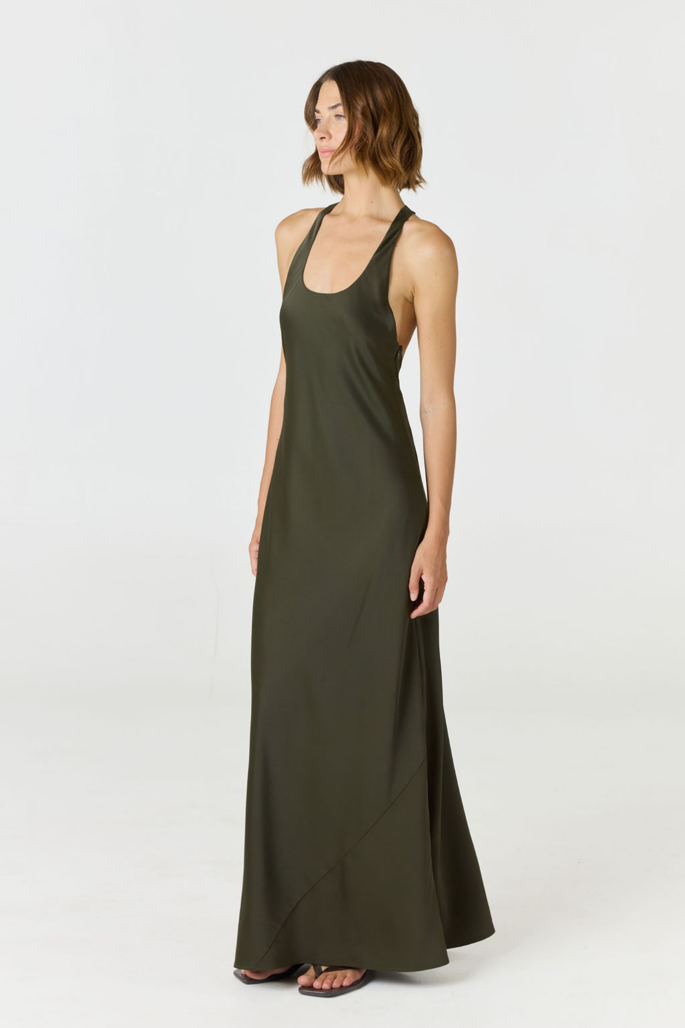 A woman modeling a sleeveless olive green maxi dress with a scoop neckline and halter-style straps, standing against a plain light gray studio background.