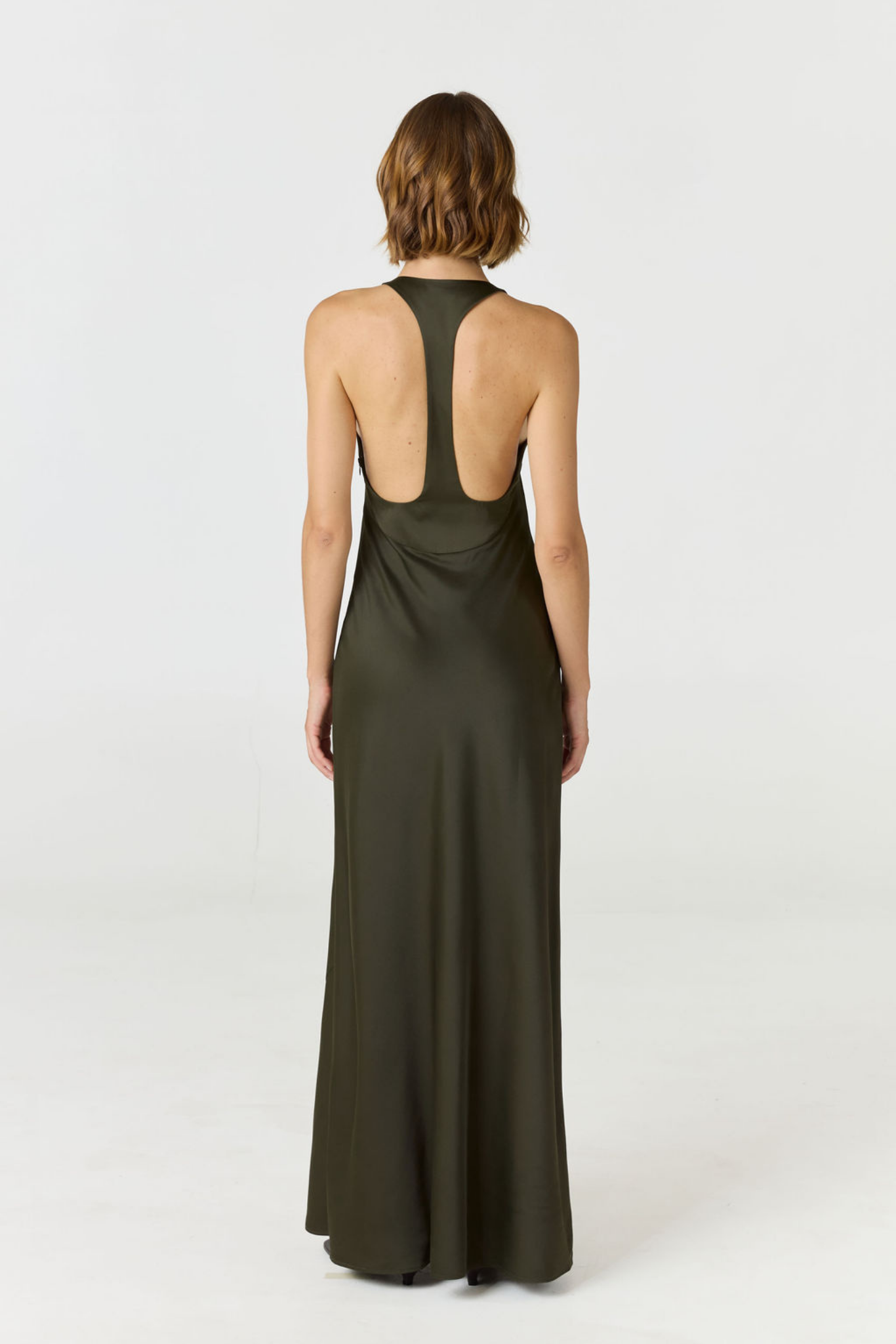 A woman viewed from the back wearing a sleeveless, floor-length dark olive green dress with a wide open back, photographed against a plain light gray studio background.