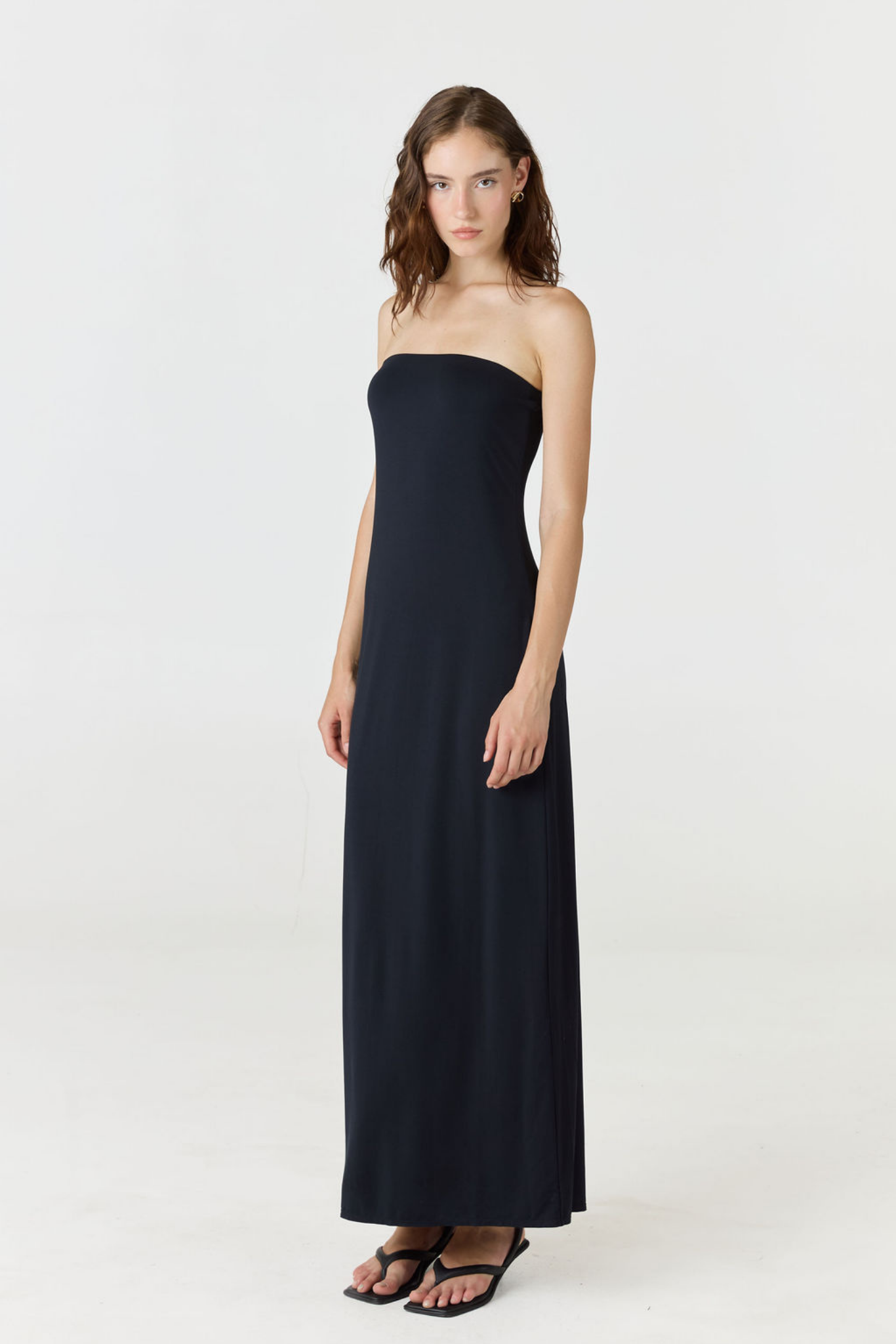A woman modeling a strapless black maxi dress with a straight neckline and a flowing, floor-length silhouette, paired with black sandals, against a plain white studio background.