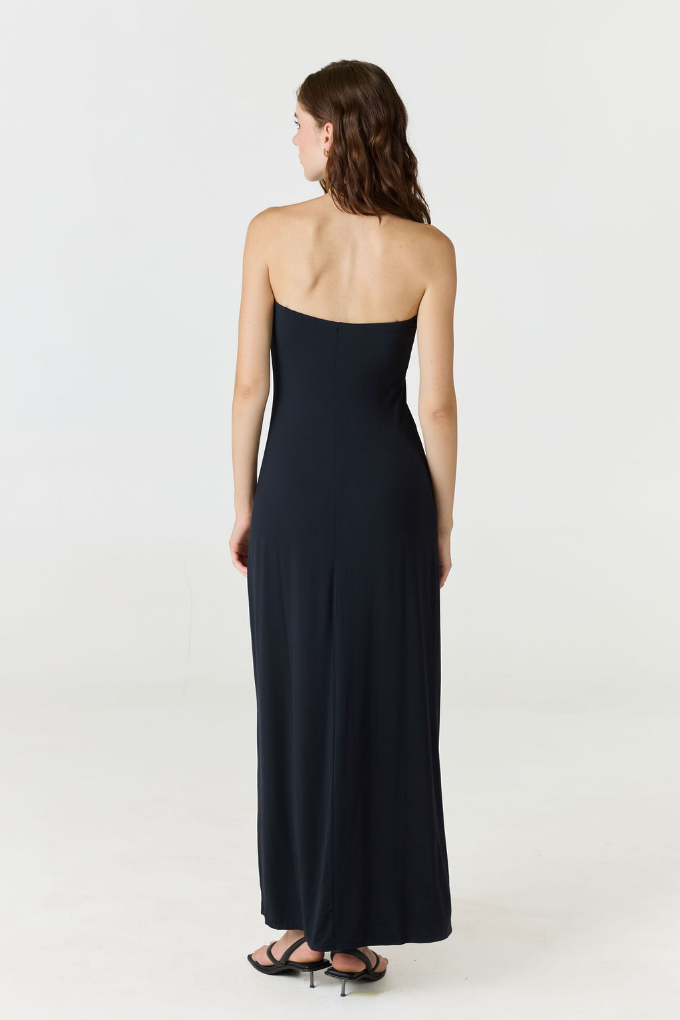 A woman viewed from the back wearing a strapless black maxi dress and black flat sandals, standing against a plain light gray studio background.