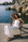 A woman wearing a white strapless jumpsuit sits in a wooden beach chair by the water, with rocky coastline and blue sea in the background.
