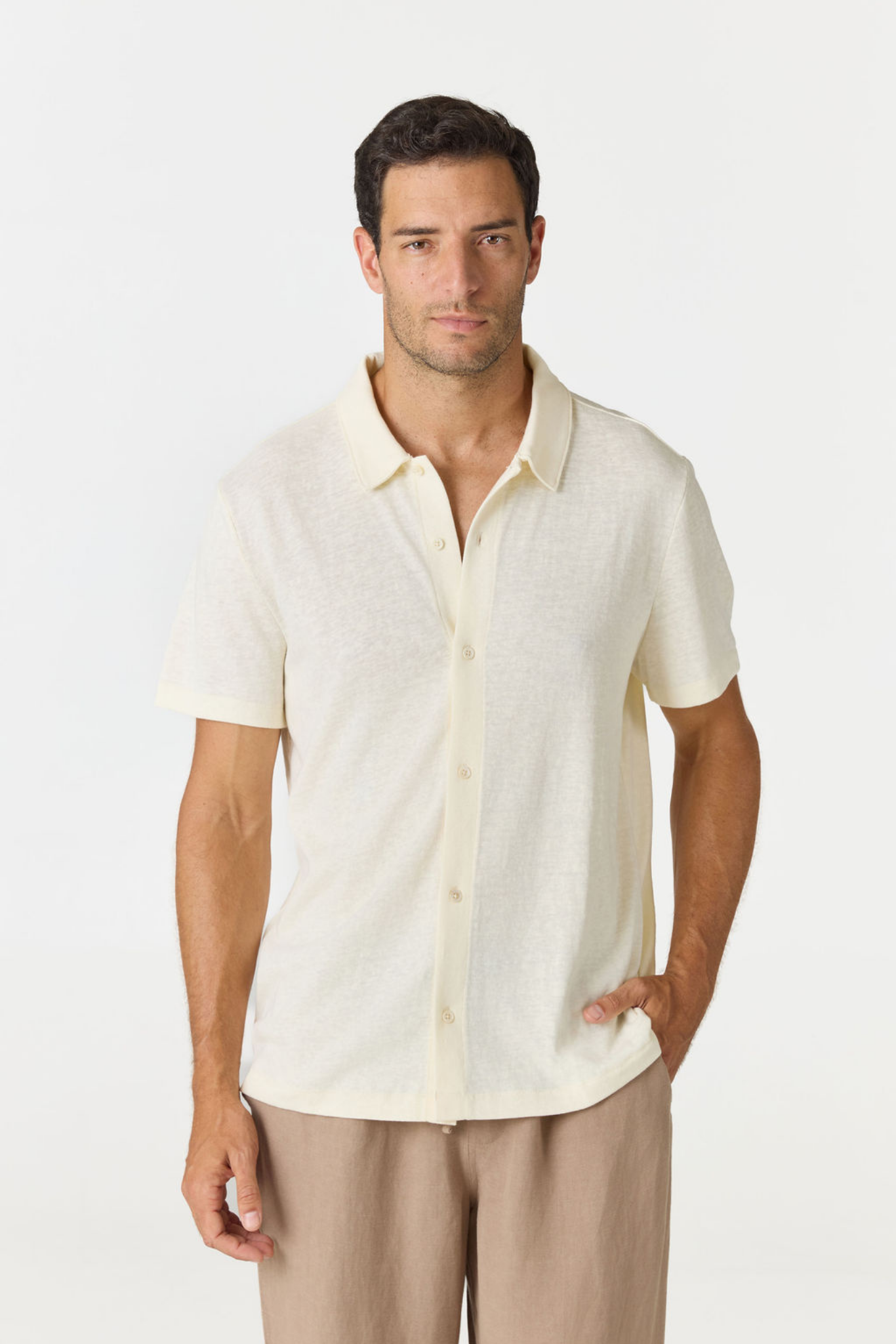 A man wearing a short-sleeve knit polo shirt in cream and tan trousers, standing against a plain light gray studio background.