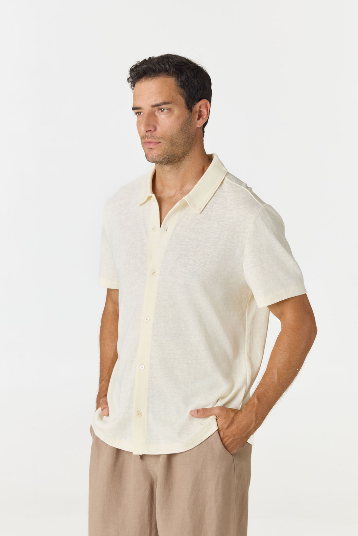A man wearing a short-sleeve knit polo shirt in a light cream color, paired with beige trousers, photographed against a plain white studio background.