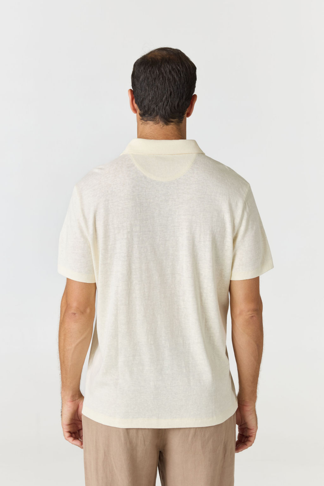 A man viewed from behind wearing a short-sleeve polo shirt in a light cream or off-white color, paired with beige trousers, against a plain light gray studio background.