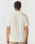 A man viewed from behind wearing a short-sleeve polo shirt in a light cream or off-white color, paired with beige trousers, against a plain light gray studio background.