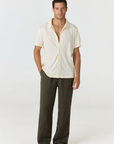 A man modeling a short-sleeve cream linen button-up shirt and dark olive green linen pants against a plain white studio background.