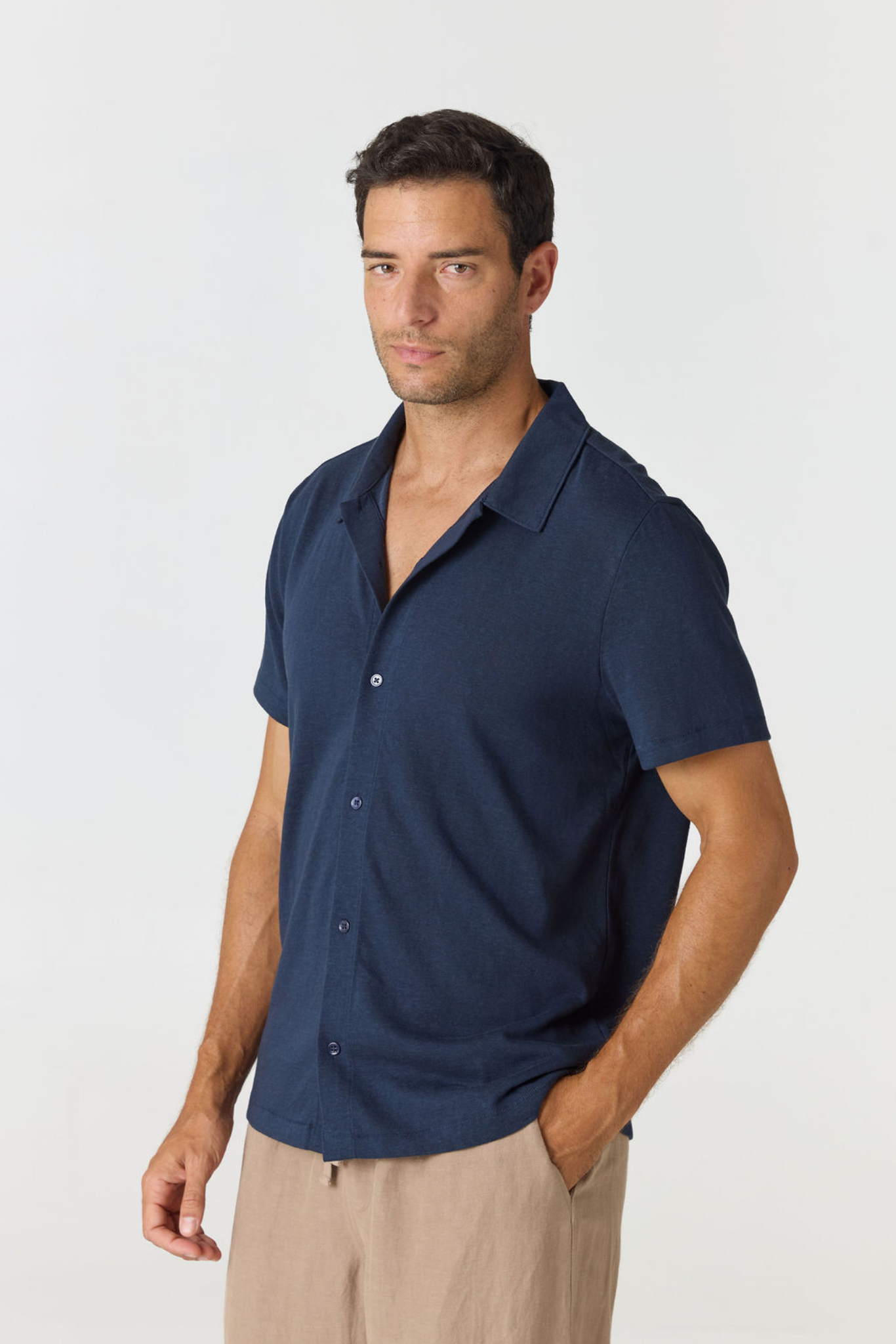 A man wearing a navy blue short-sleeve button-up shirt and beige trousers, standing against a light gray studio background.
