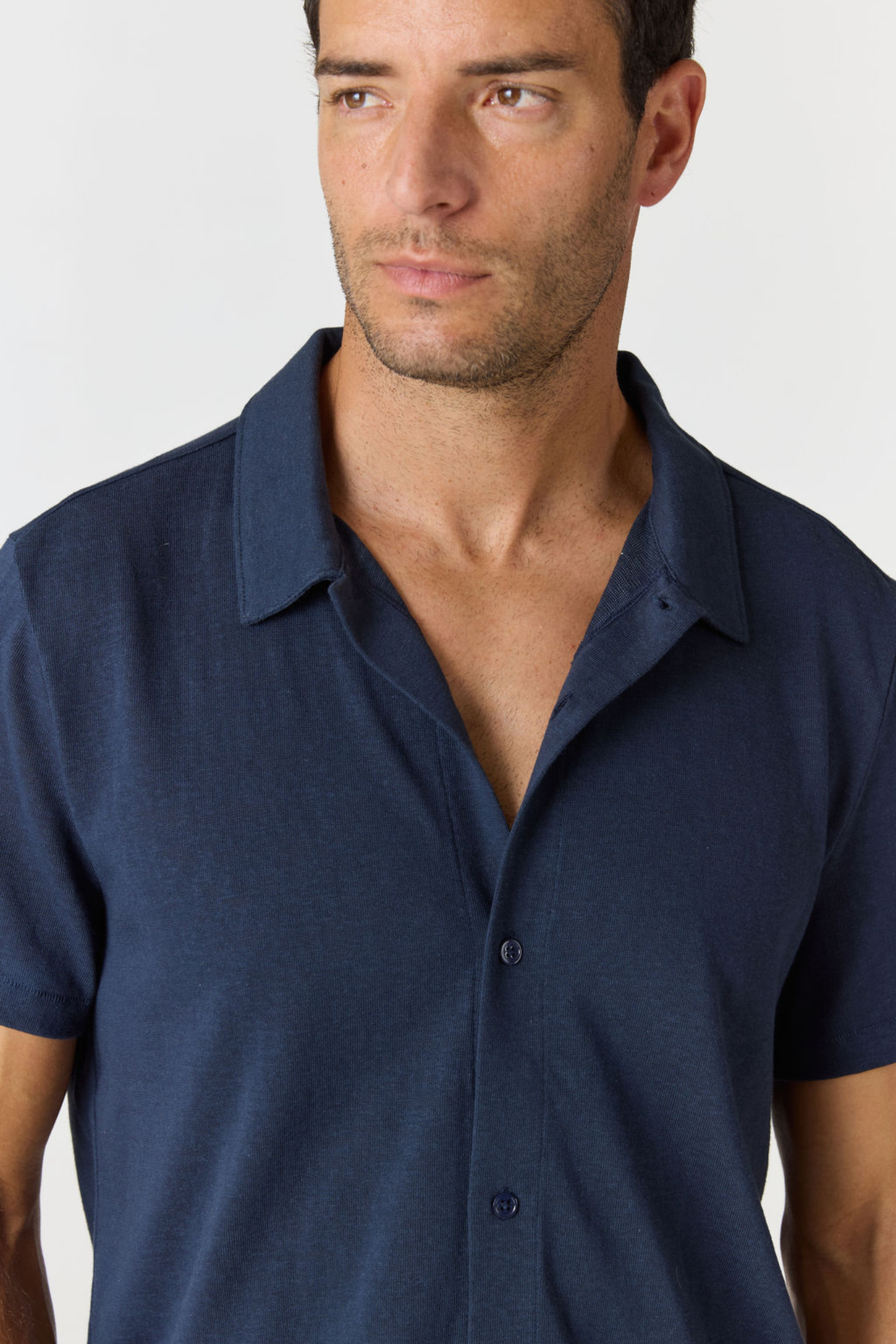 A close-up of a man wearing a dark navy blue short-sleeve button-up shirt, cropped from the chest up against a light gray background.