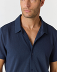 A close-up of a man wearing a dark navy blue short-sleeve button-up shirt, cropped from the chest up against a light gray background.