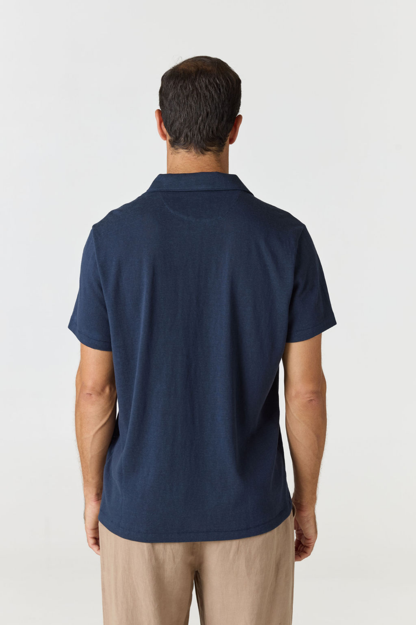 A man viewed from the back wearing a navy blue short-sleeve polo shirt and beige trousers, standing against a plain light gray studio background.