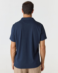A man viewed from the back wearing a navy blue short-sleeve polo shirt and beige trousers, standing against a plain light gray studio background.