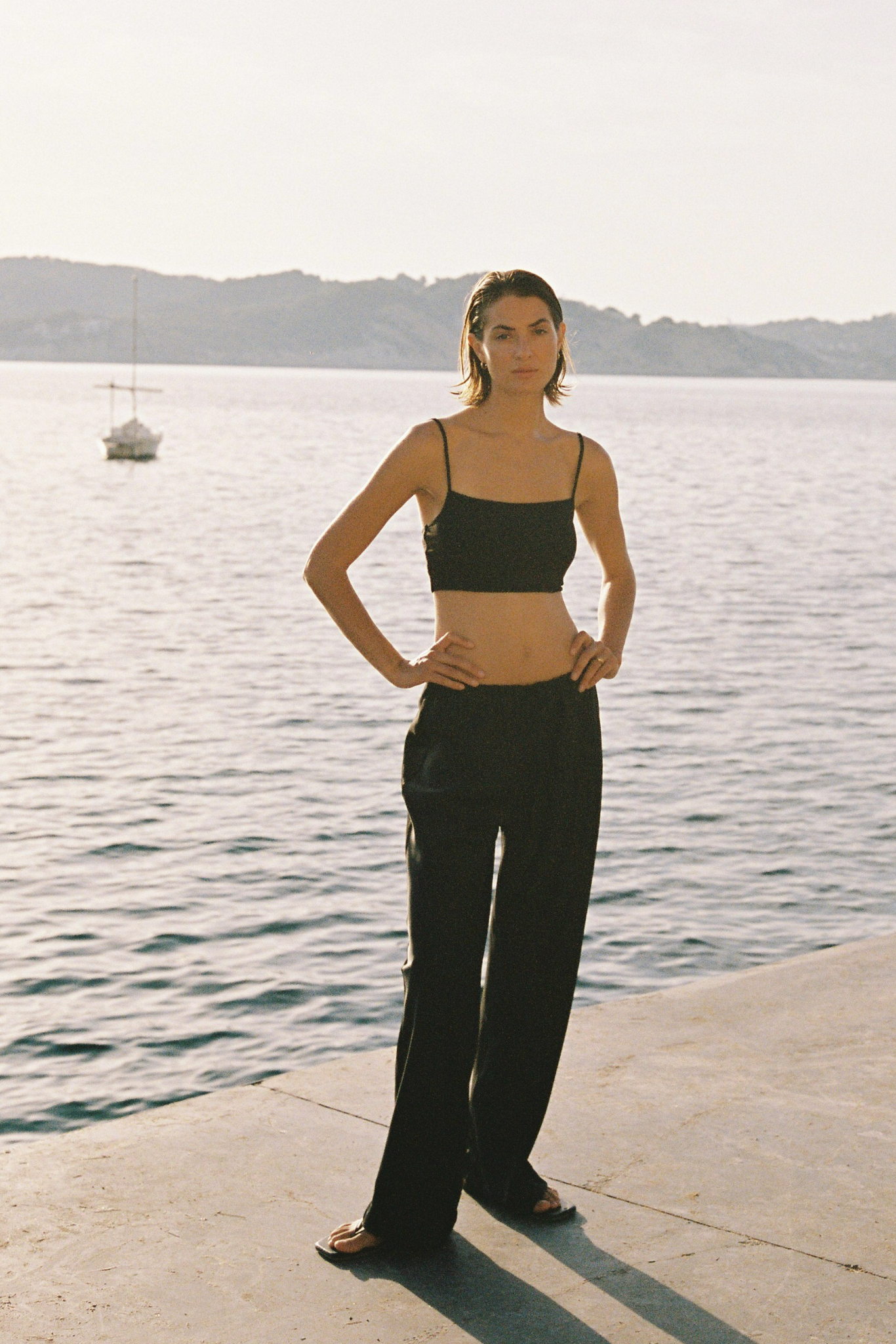 A woman stands on a concrete pier by the water wearing a black spaghetti-strap crop top and matching black wide-leg trousers, with a sailboat and distant shoreline in the background.