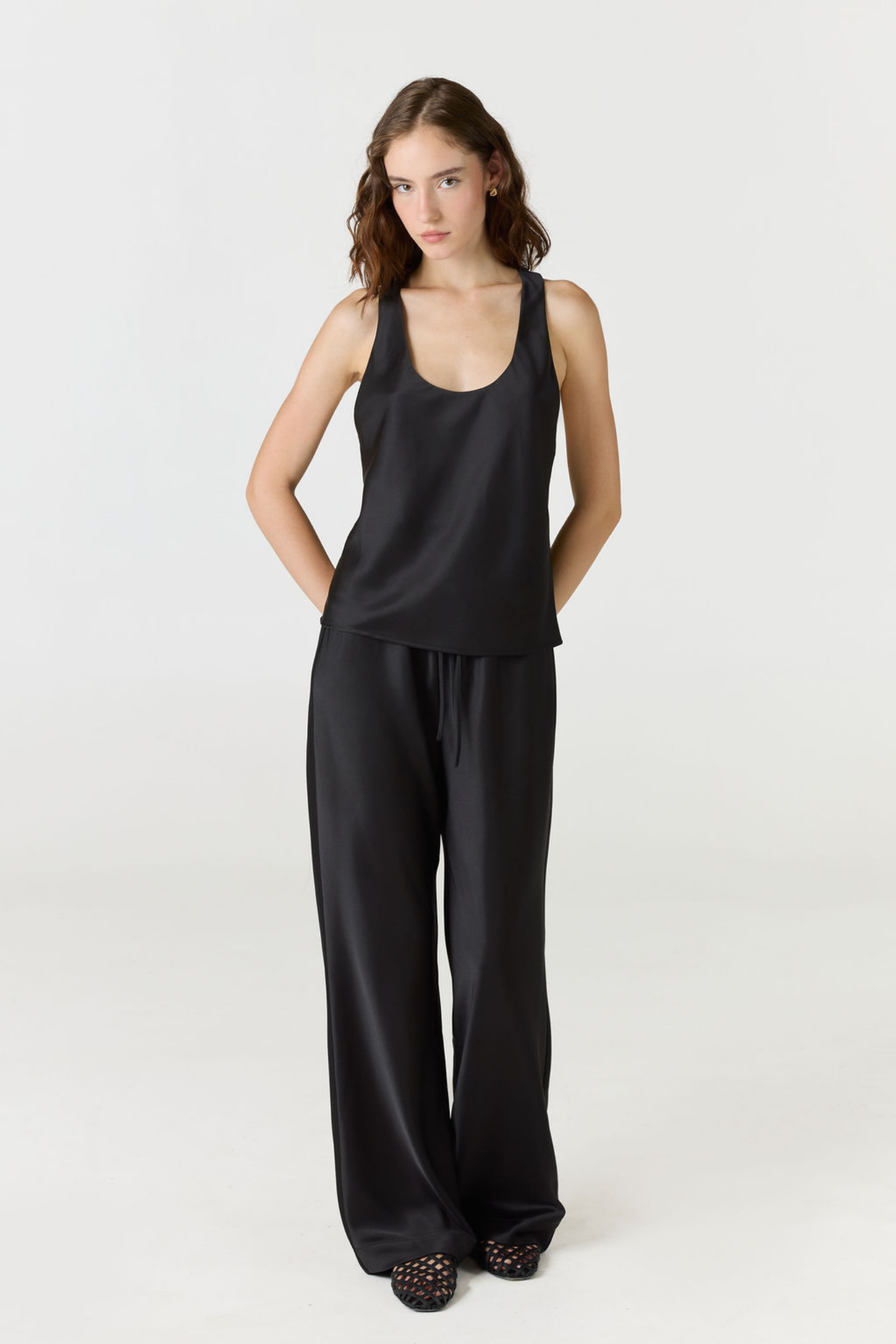 A woman modeling a black satin-like sleeveless top and matching wide-leg pants against a plain light gray studio background.