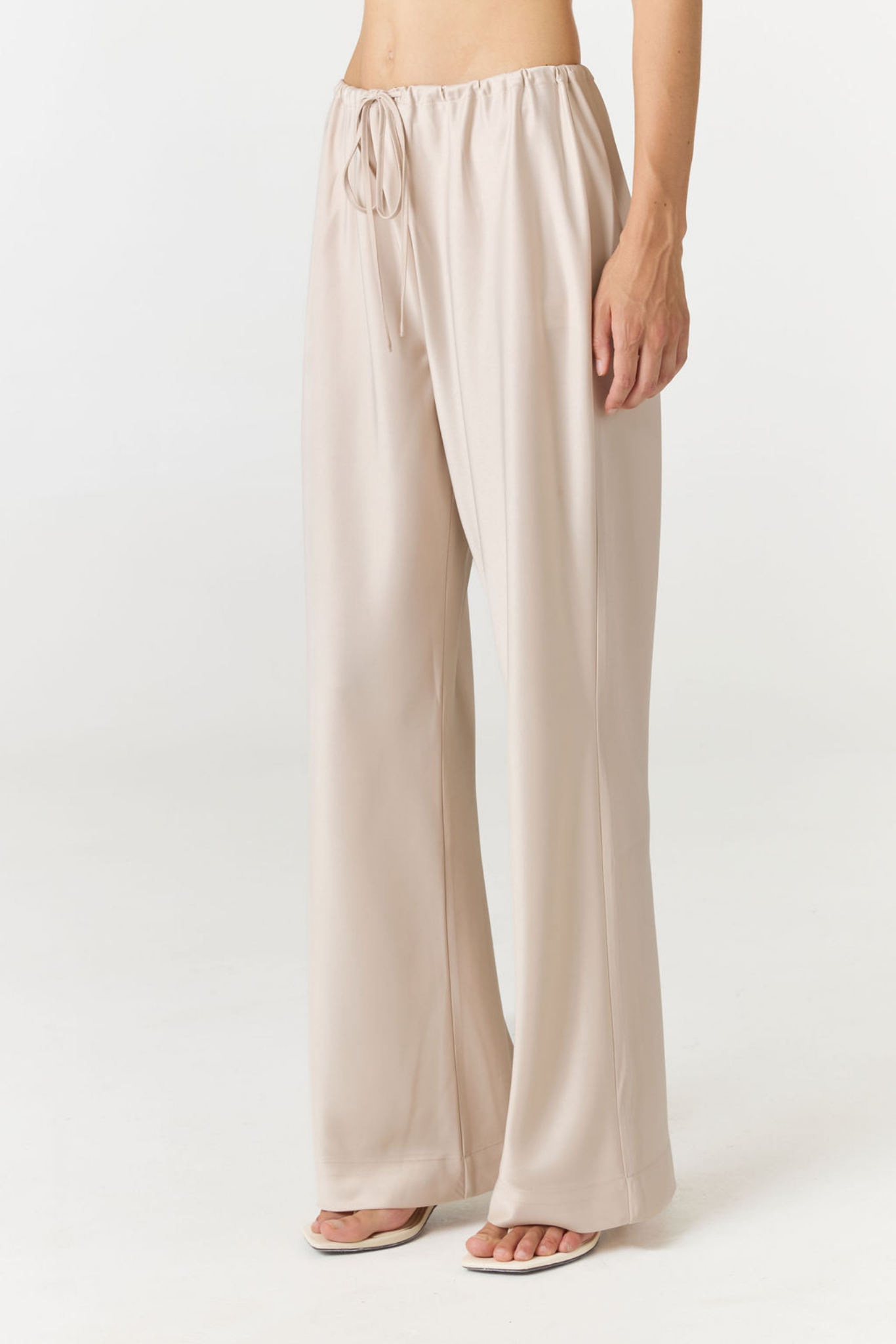 A model wearing light beige wide-leg drawstring pants and white slide sandals, shown from the waist down against a plain white studio background.