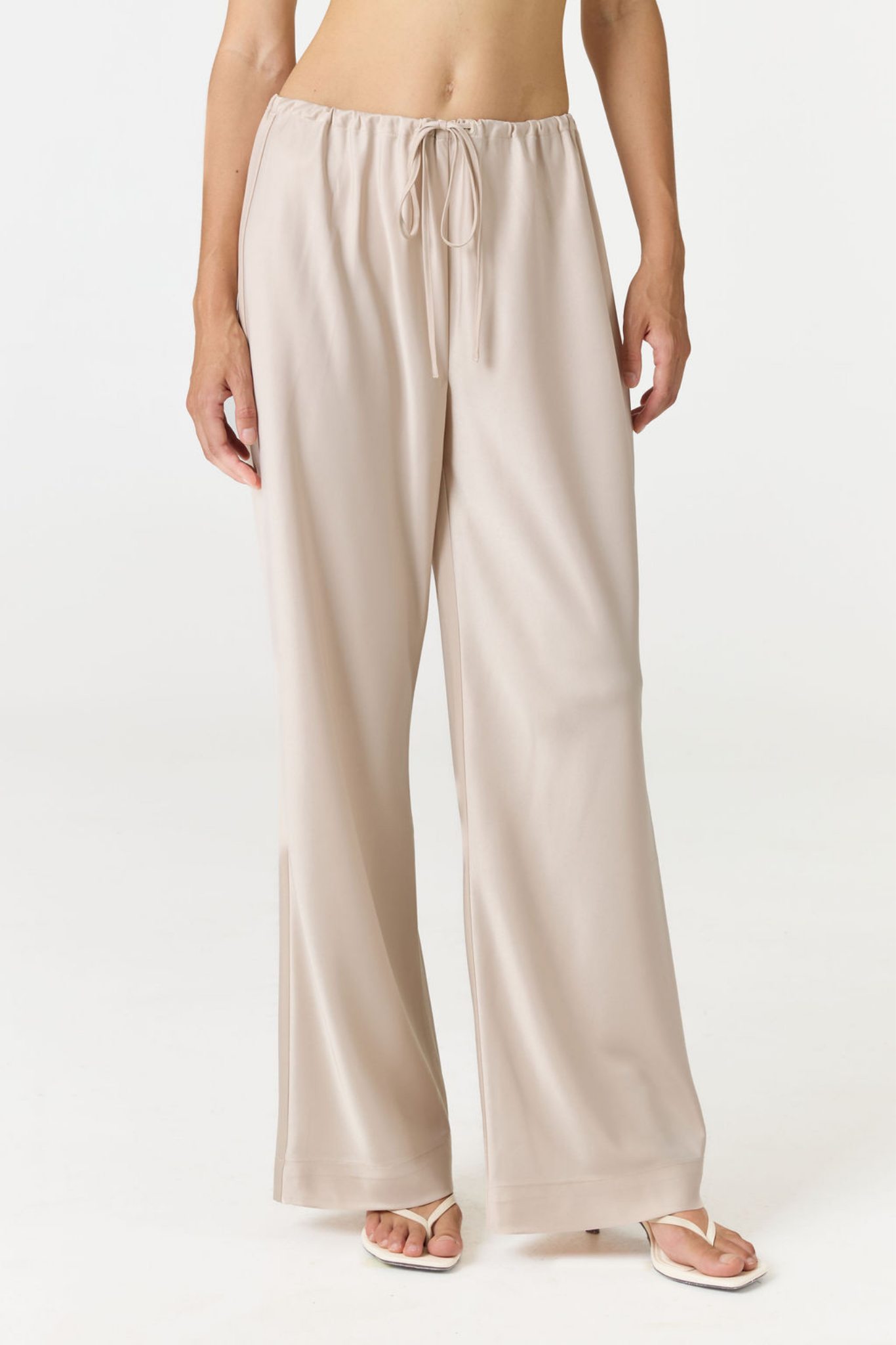 A model wearing light beige wide-leg lounge pants with a drawstring waistband, paired with white thong sandals, shown from the waist down against a plain white studio background.