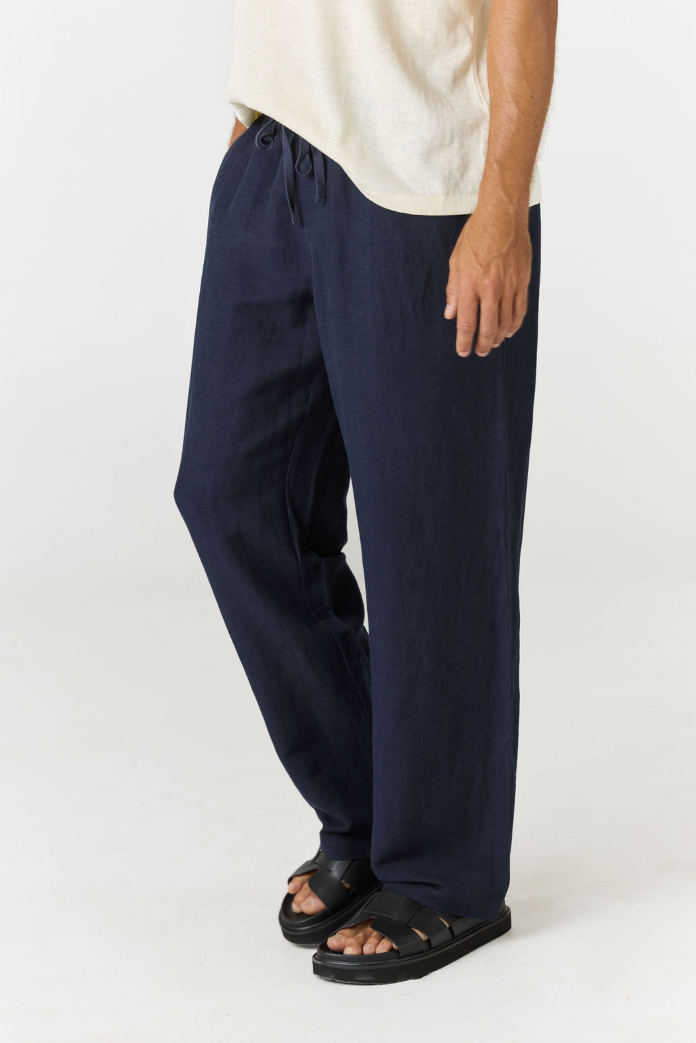 A person wearing loose, wide-leg navy pants with a drawstring waist, paired with a light cream t-shirt and black slide sandals, shown from the waist down on a plain white studio background.
