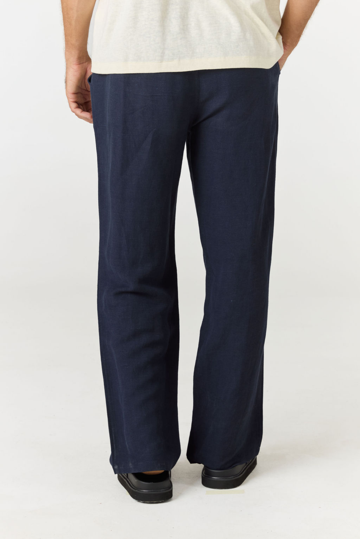 A person shown from the back wearing dark navy wide-leg trousers, a light cream top, and black shoes against a plain light gray studio background.