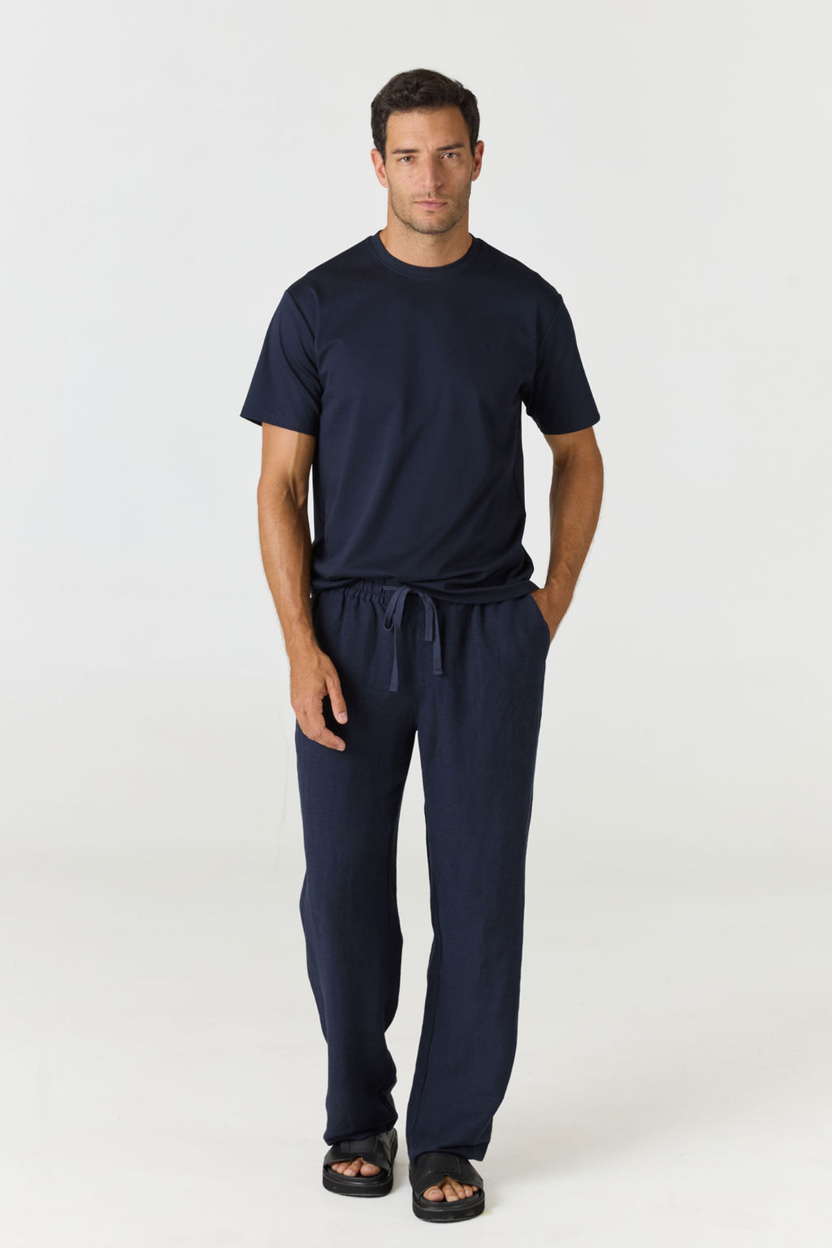 A man modeling a navy blue scrub set consisting of a short-sleeve crewneck shirt and matching drawstring pants, paired with black slide sandals, against a plain white studio background.
