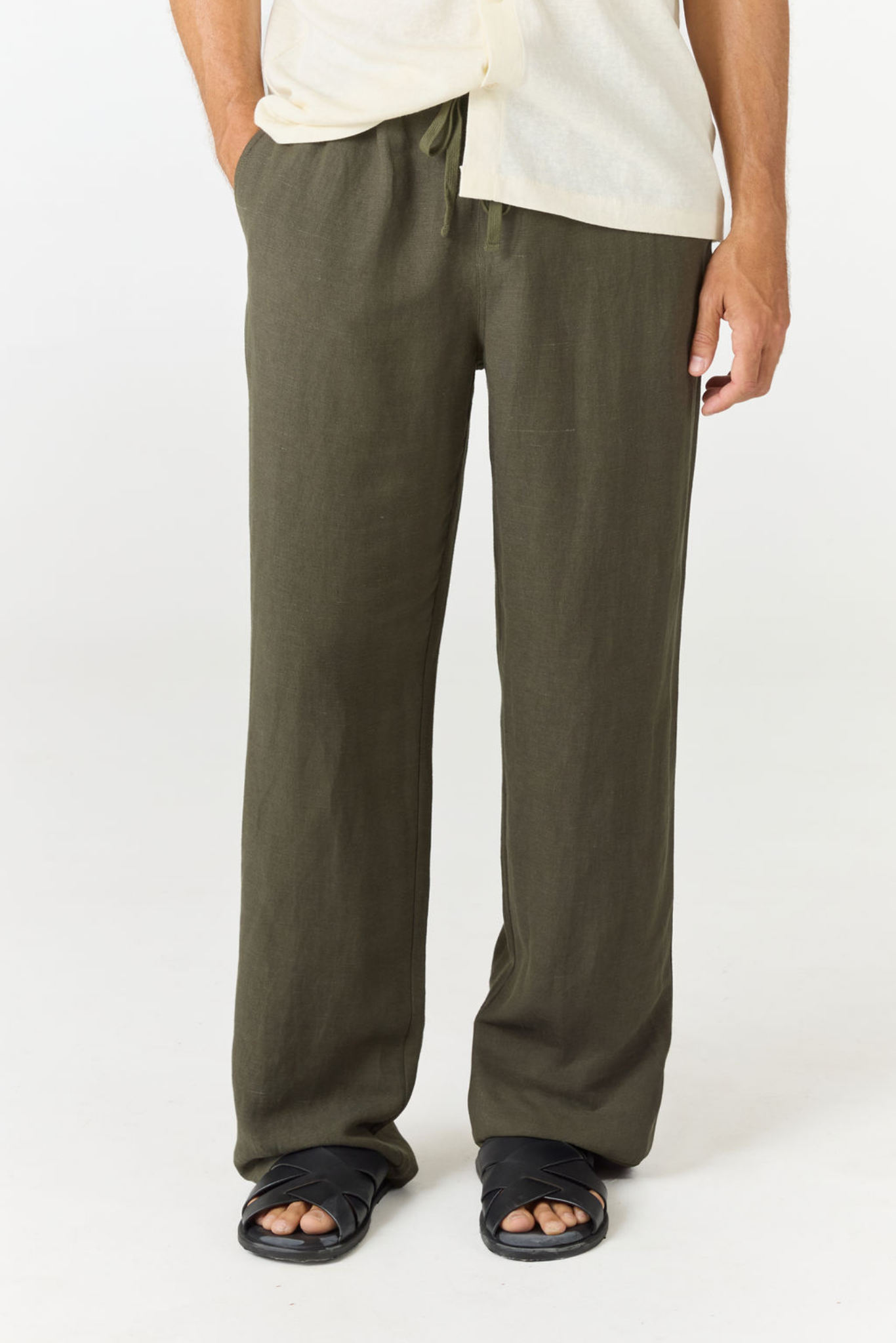 A person wearing loose, wide-leg olive green pants with a cream shirt and black sandals, shown from the waist down against a plain white studio background.