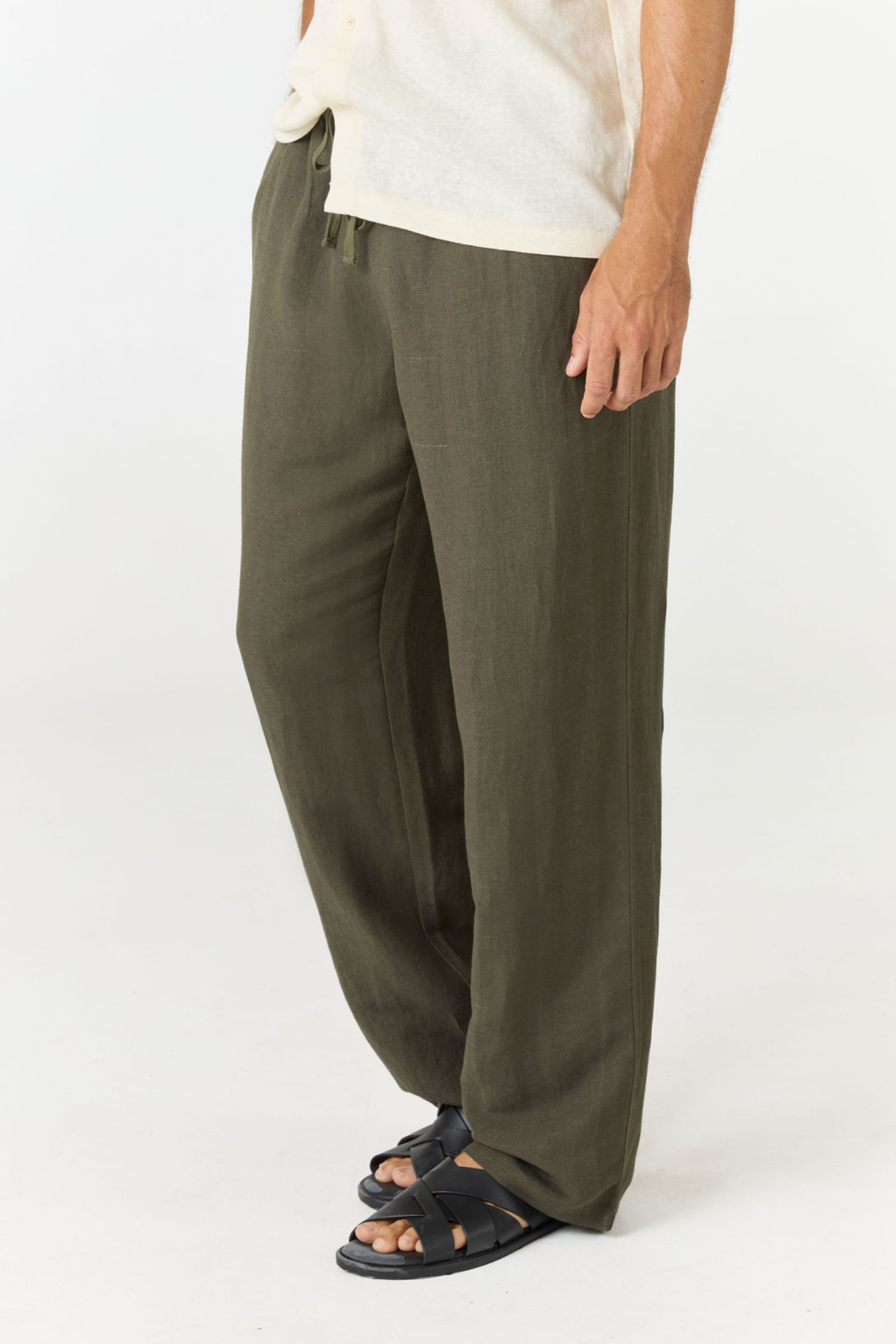 A person wearing loose, wide-leg olive green pants with a drawstring waist, paired with a light cream shirt and black sandals, shown against a plain white studio background.