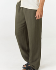 A person wearing loose, wide-leg olive green pants with a drawstring waist, paired with a light cream shirt and black sandals, shown against a plain white studio background.