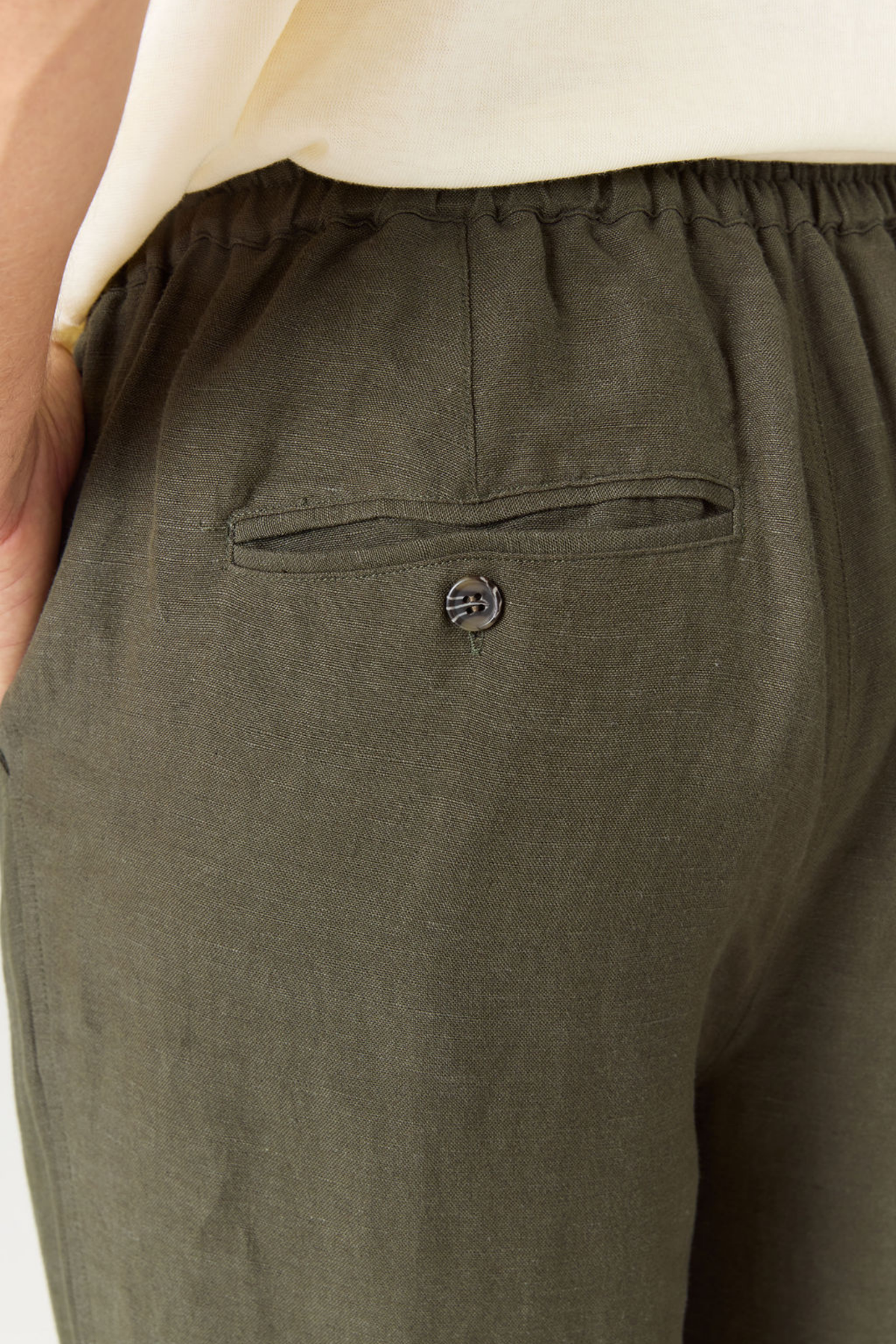Close-up of olive green linen trousers with an elastic waistband and a side welt pocket featuring a decorative metal button.