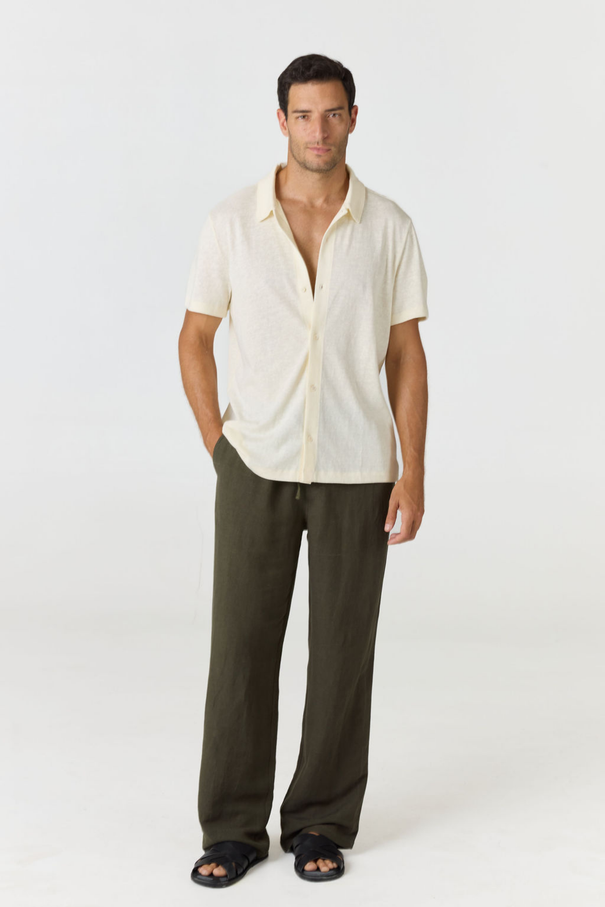 A man modeling a short-sleeve cream linen button-up shirt and dark olive green linen pants against a plain white studio background.