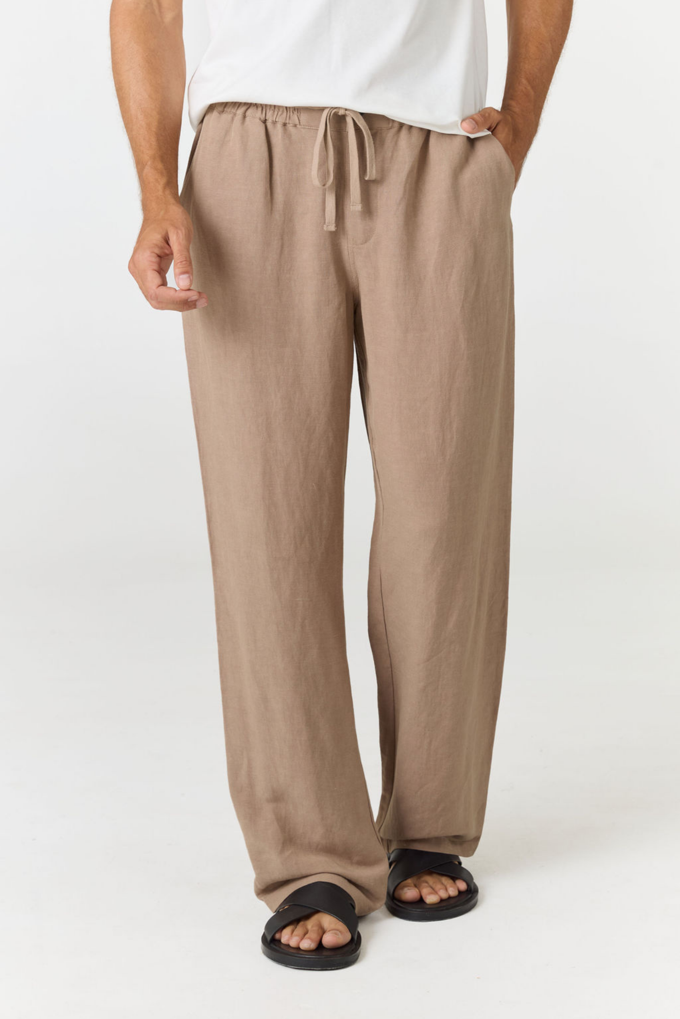 A person wearing loose, wide-leg taupe pants with a drawstring waist, paired with a white top and black sandals, shown from the waist down against a plain light background.