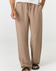 A person wearing loose, wide-leg taupe pants with a drawstring waist, paired with a white top and black sandals, shown from the waist down against a plain light background.