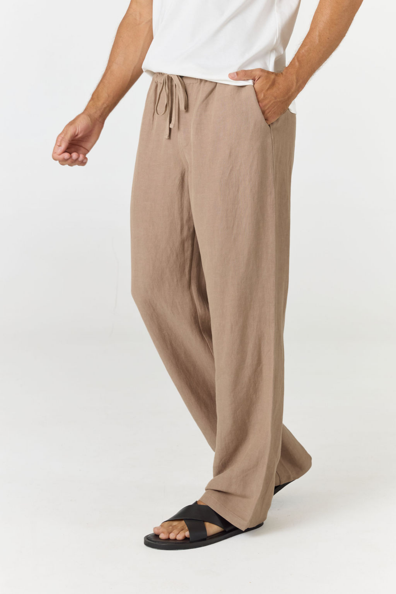 A person wearing loose, wide-leg taupe pants with a drawstring waist, paired with a white top and black slide sandals, shown against a plain light gray studio background.