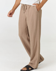 A person wearing loose, wide-leg taupe pants with a drawstring waist, paired with a white top and black slide sandals, shown against a plain light gray studio background.