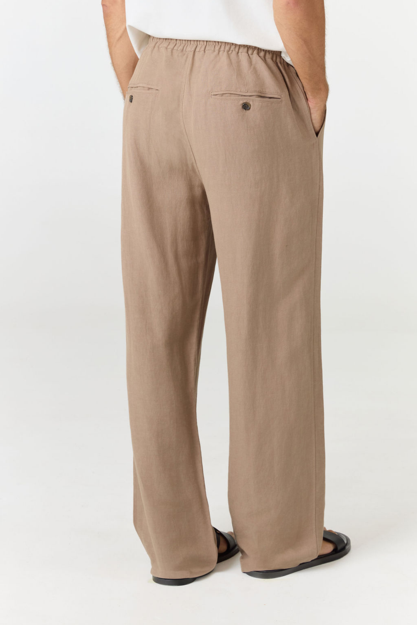 A person shown from the back wearing loose, wide-leg taupe pants with an elastic waistband, side pockets, and a small back button, paired with a white top and black sandals on a light gray studio background.