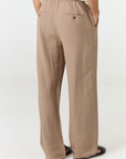 A person shown from the back wearing loose, wide-leg taupe pants with an elastic waistband, side pockets, and a small back button, paired with a white top and black sandals on a light gray studio background.