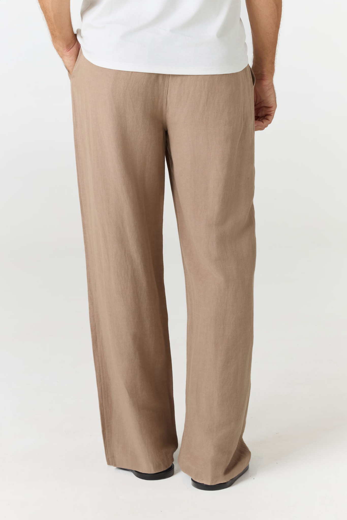 A person wearing loose, wide-leg taupe pants with a white top, shown from the back against a plain light gray studio background.