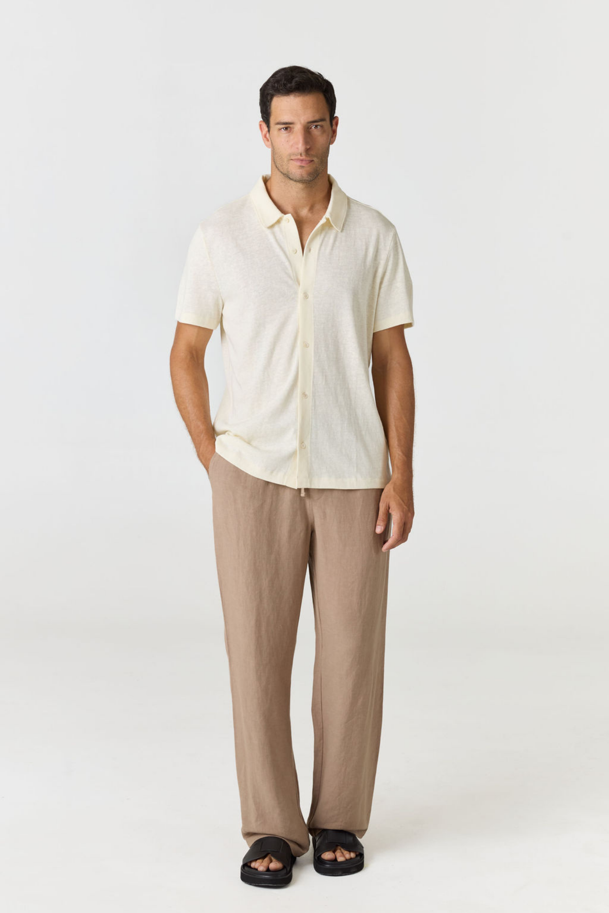 A man modeling a short-sleeve cream linen button-up shirt and beige linen trousers with black slide sandals, photographed against a seamless light gray studio background.