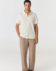 A man modeling a short-sleeve cream linen button-up shirt and beige linen trousers with black slide sandals, photographed against a seamless light gray studio background.
