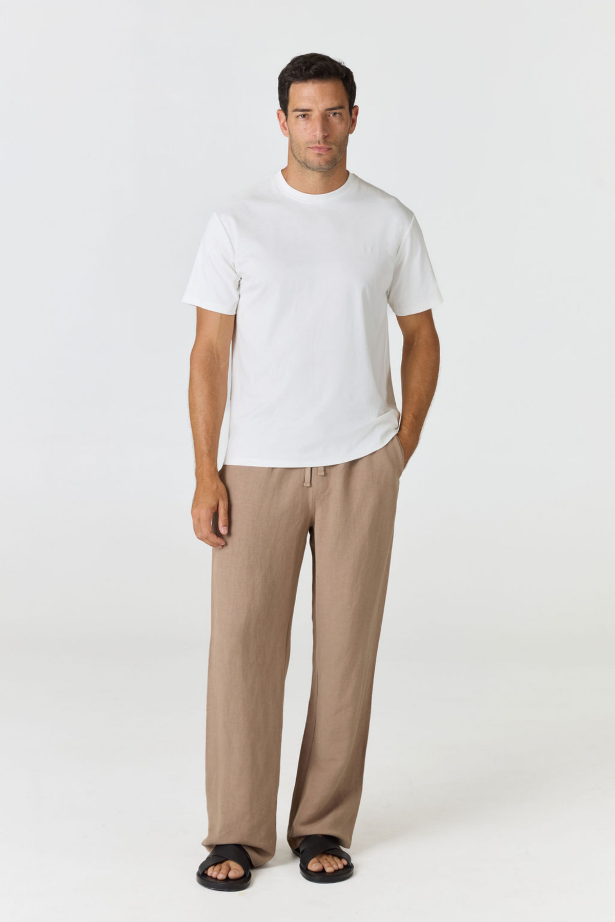 A man modeling a white short-sleeve crewneck t-shirt and beige wide-leg trousers, standing against a plain light gray studio background.
