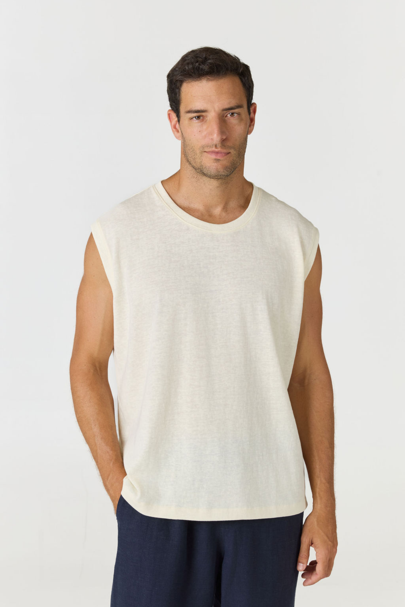 A man wearing a sleeveless crew-neck knit top in off-white, paired with navy pants, photographed against a light gray studio background.