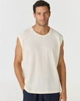 A man wearing a sleeveless crew-neck knit top in off-white, paired with navy pants, photographed against a light gray studio background.