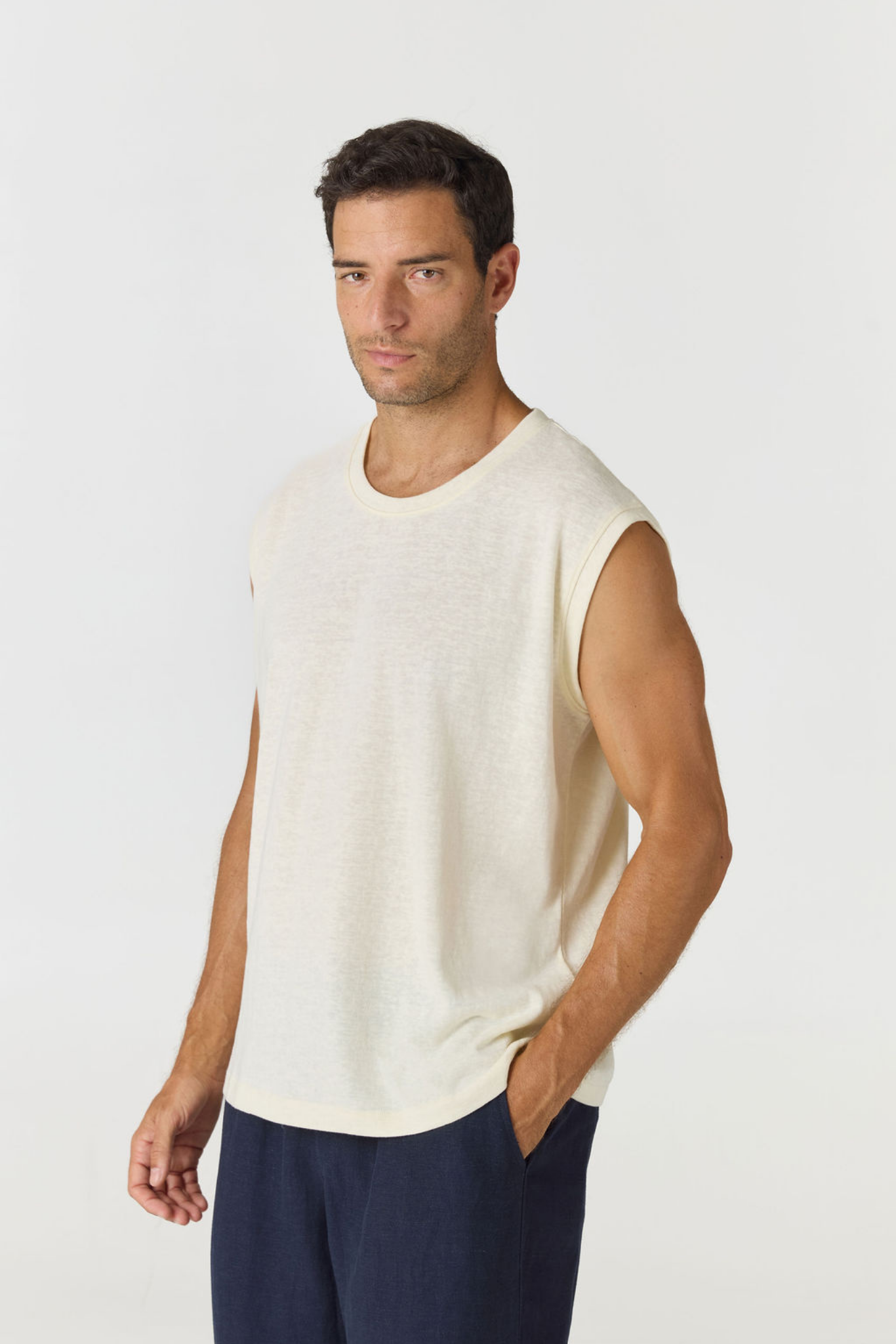 A man wearing a sleeveless cream-colored knit top and navy blue pants, standing against a light gray studio background.