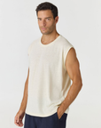 A man wearing a sleeveless cream-colored knit top and navy blue pants, standing against a light gray studio background.