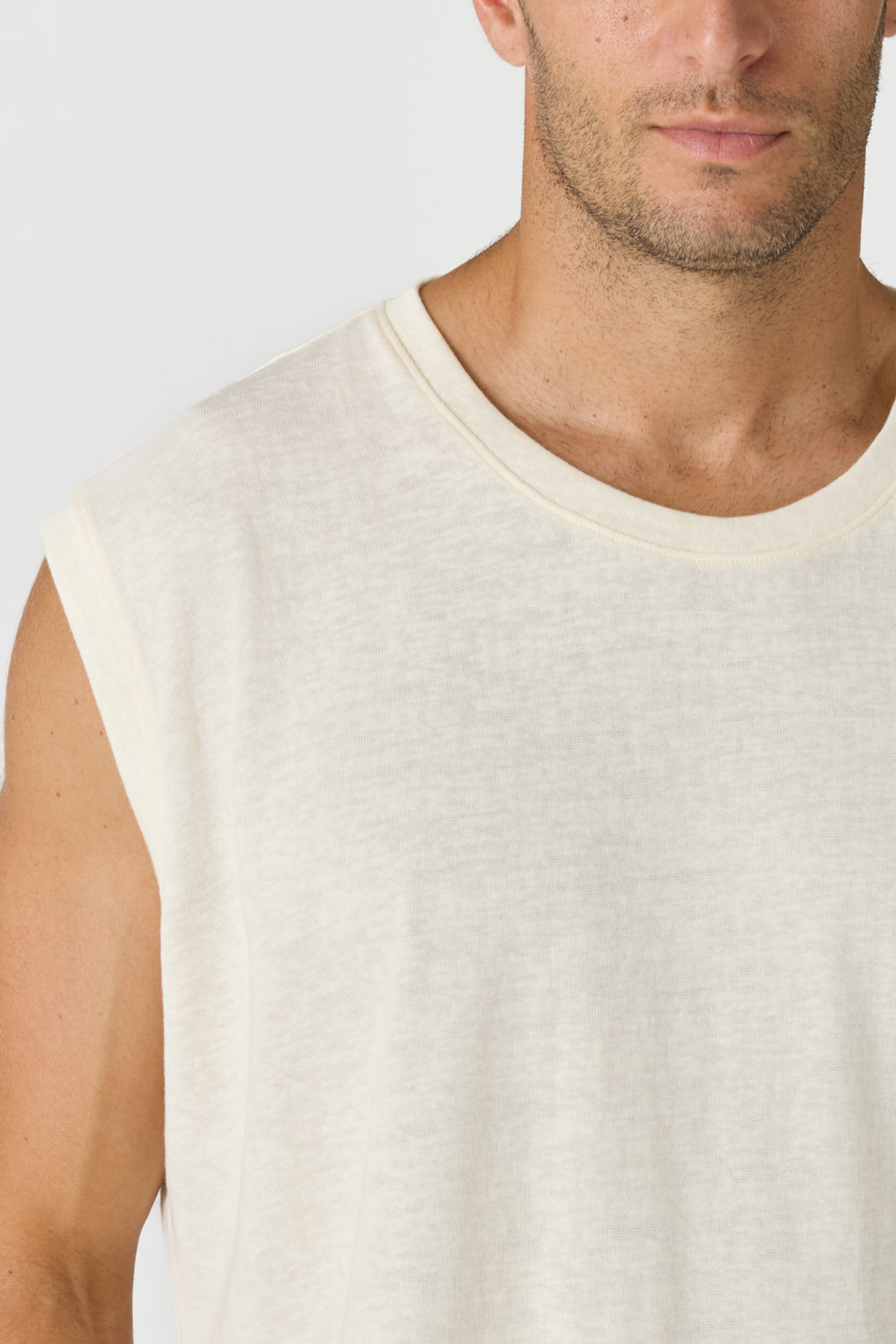 Close-up of a man wearing a sleeveless crew-neck t-shirt in a light cream or off-white color, showing the fabric texture and fit.